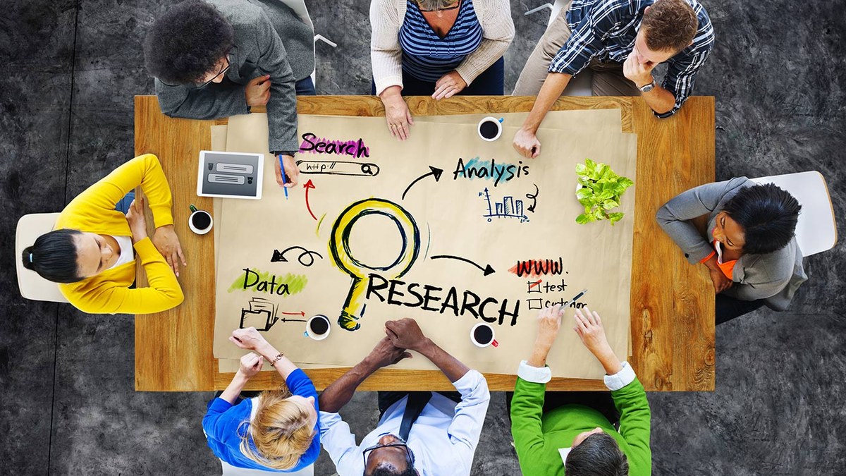 Top-down view of a diverse team brainstorming around a table with a large 'RESEARCH' diagram and ideas like data, search, and analysis drawn on paper.
