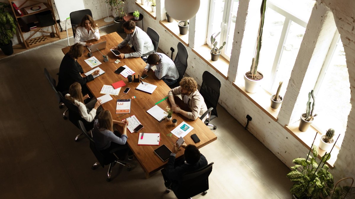Eight colleagues sit around a long wooden conference table in a bright, plant-filled office, collaborating with papers and laptops on the table,