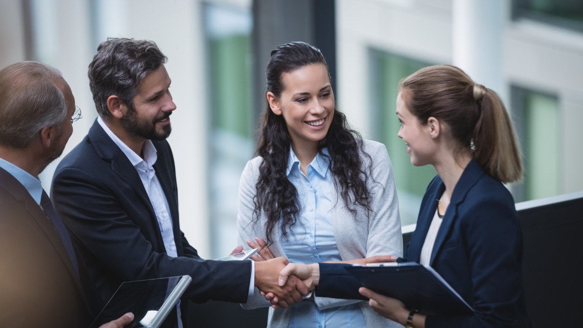 Four professionals in business attire shake hands and smile in a modern office lobby. 2 men and 2 women present, one holds a tablet.