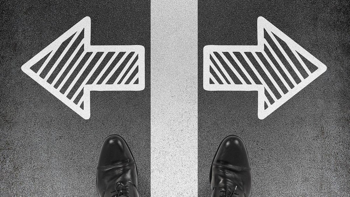 Person wearing black shoes stands at a fork in the road, arrows pointing left and right ahead of them.”