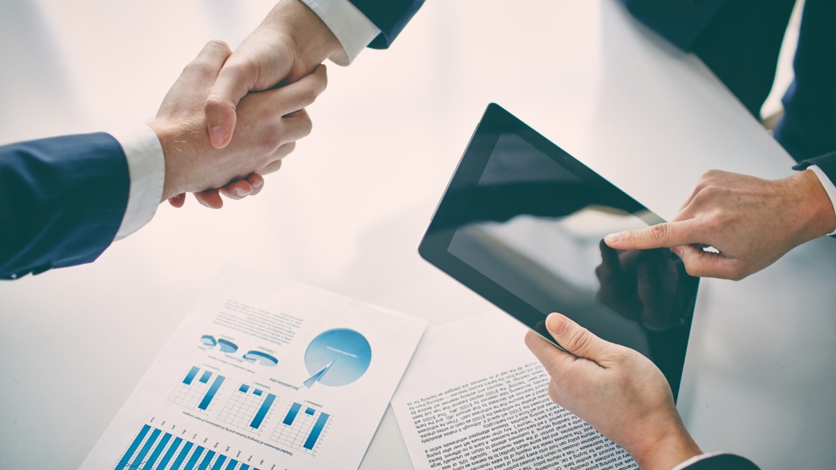 Two professionals shake hands over a table with printed charts and a tablet nearby.