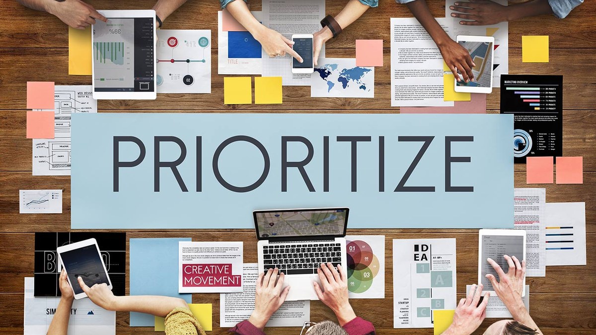 Top-down view of a team workspace with a large blue 'PRIORITIZE' sign amid laptops, tablets, and scattered documents.