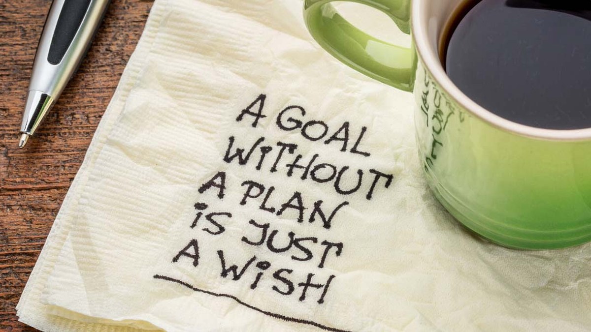 Napkin with a handwritten quote: 'A goal without a plan is just a wish,' beside a pen and a green coffee mug on a wooden table.