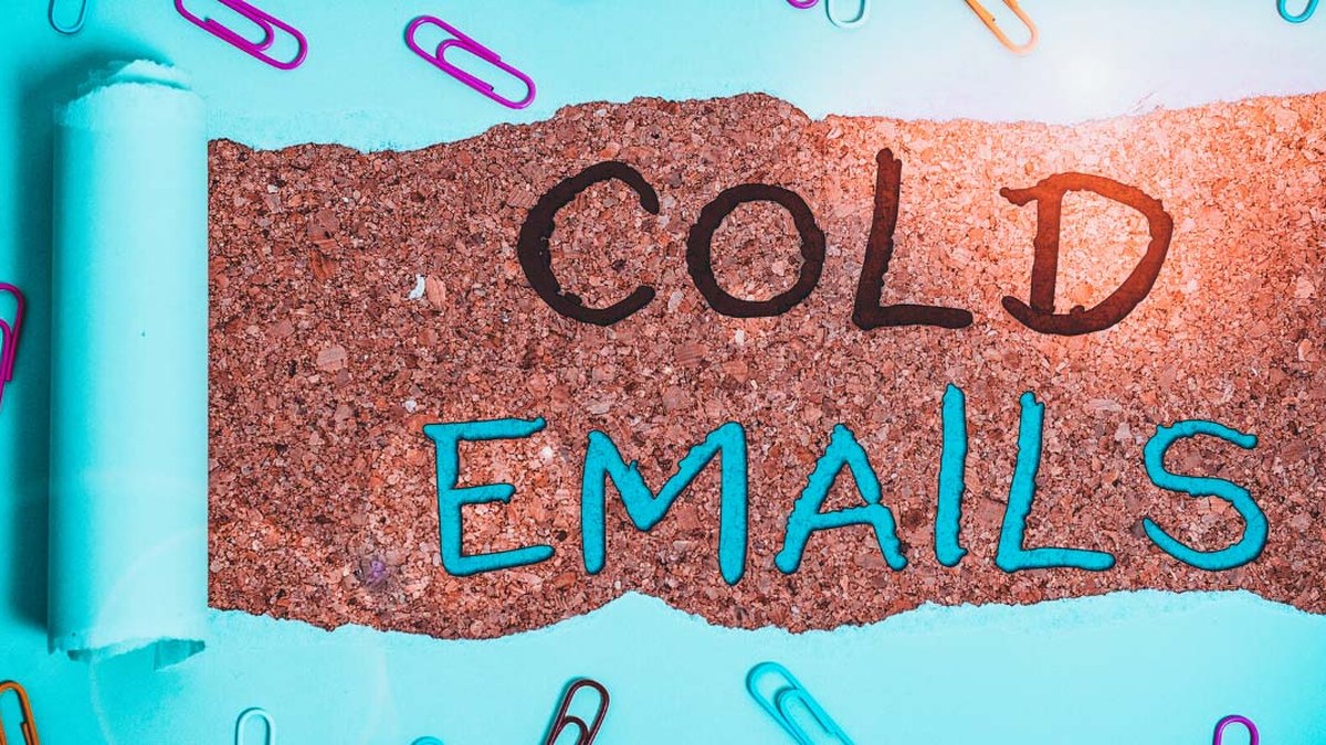 Cork strip with the words 'COLD EMAILS' written in black and turquoise on a teal background with colorful paperclips nearby.