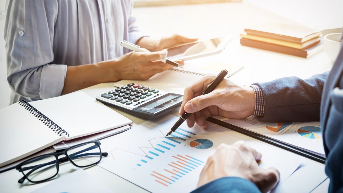 Two professionals analyzing financial charts at a desk, with a calculator, notebook, and glasses nearby.