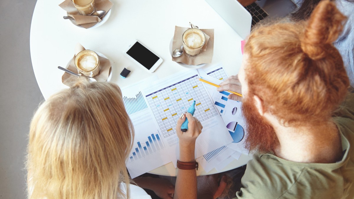 Overhead view of two people analyzing printed charts and graphs at a round table with coffee cups and a smartphone nearby.