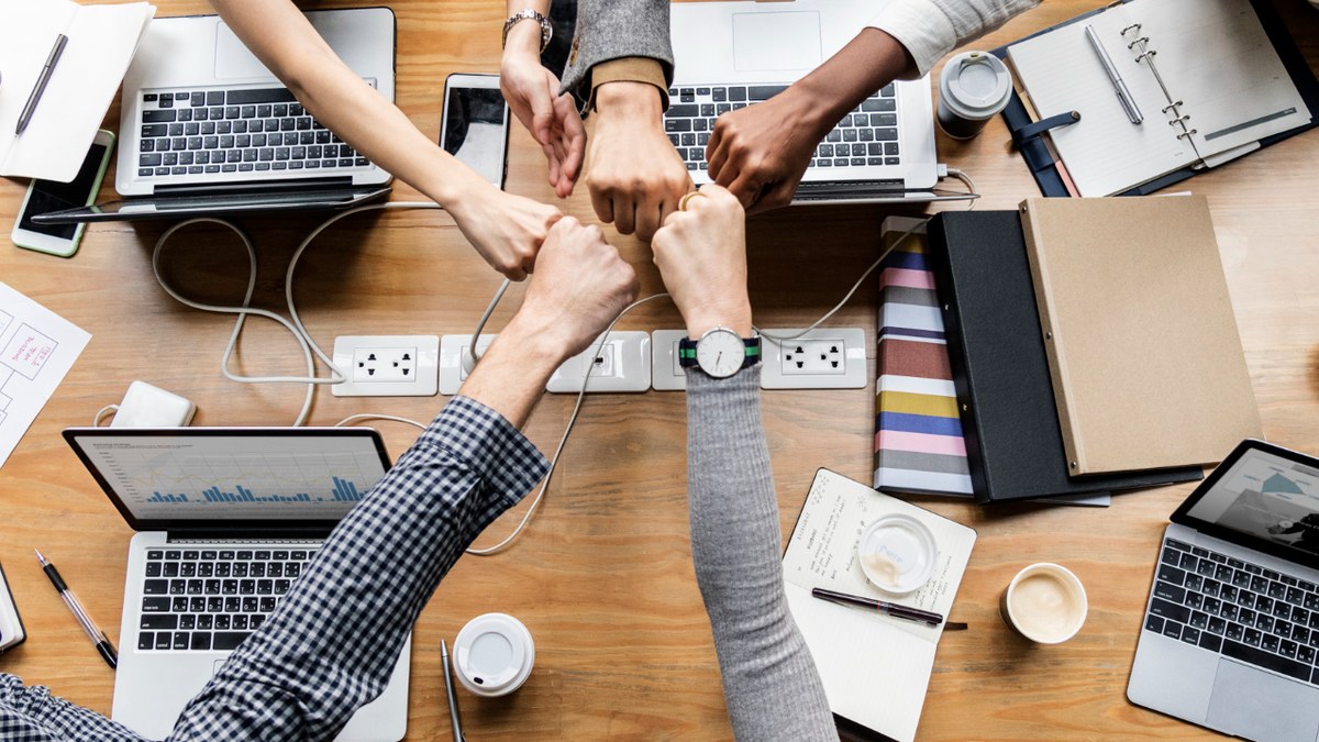 Team of coworkers performing a fist bump over a wooden table with laptops, notebooks, and coffee cups, symbolizing teamwork and collaboration.