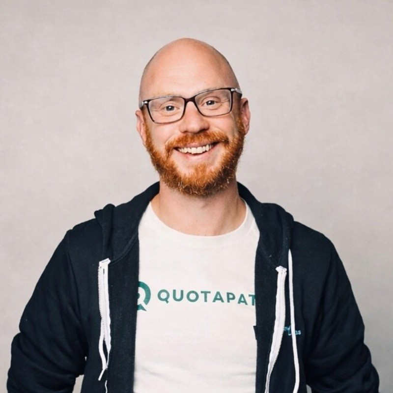 Portrait of a smiling man with glasses, a red beard, and a bald head, wearing a white t-shirt and black zip-up hoodie against a light gray background.